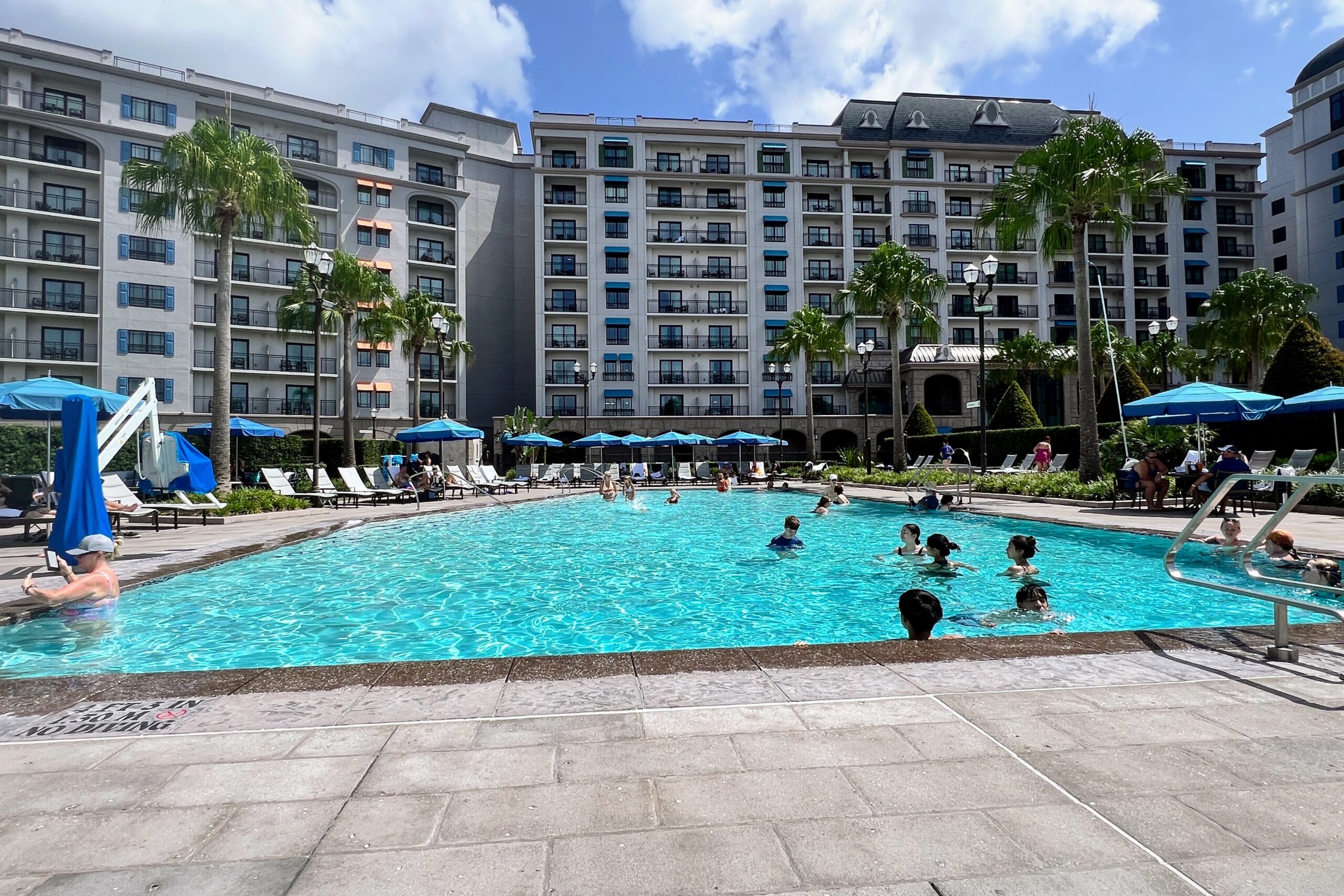 People swim and relax at a resort pool with palm trees and hotels in the background.