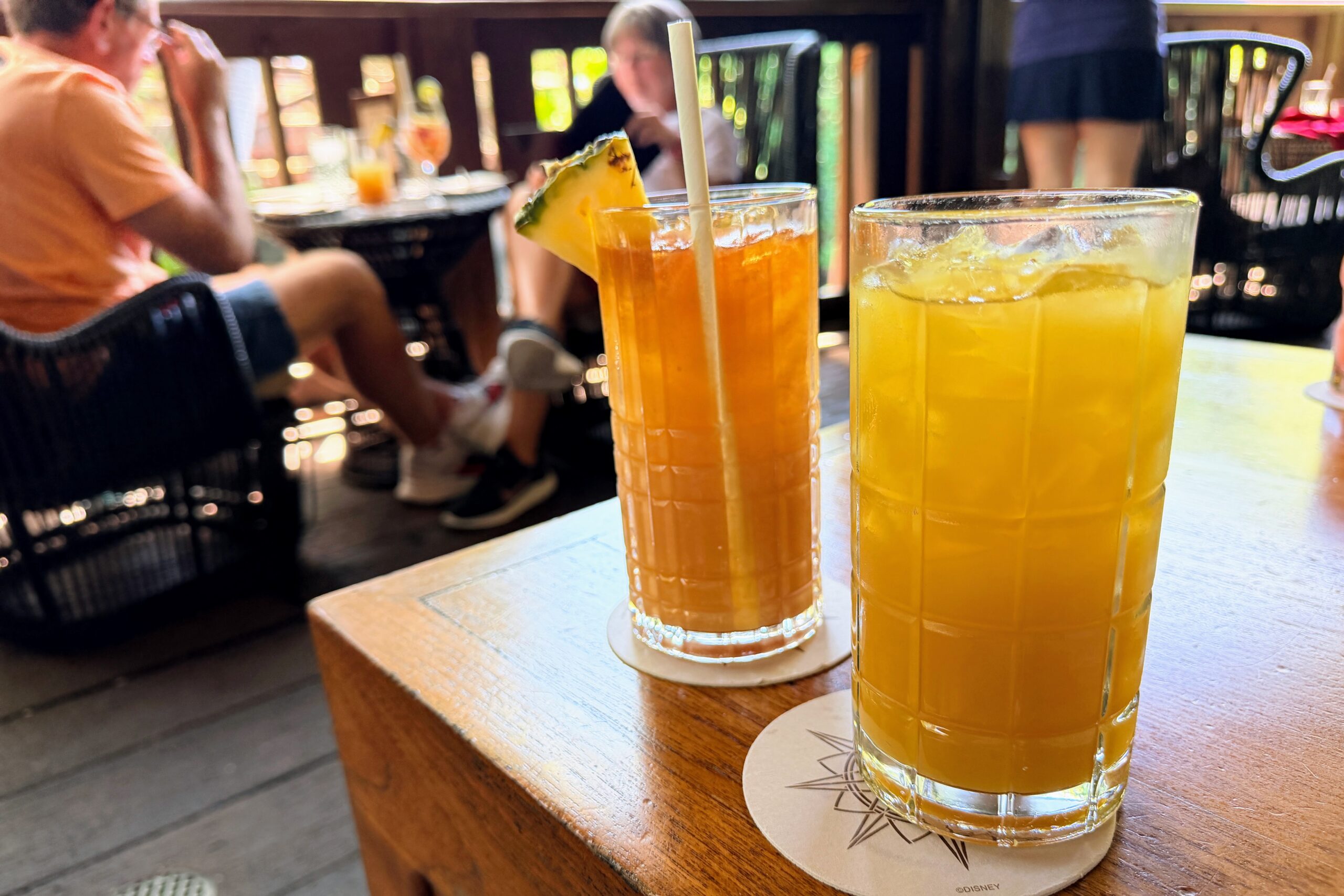 Two tropical drinks with pineapple on a wooden deck, people in background
